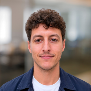 Close-up portrait of a young man with curly brown hair, wearing a navy jacket and white t-shirt, smiling softly at the camera in a blurred office setting.