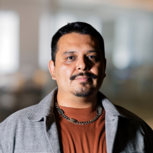 Head-and-shoulders portrait of a man with short black hair and a goatee, wearing an orange-brown shirt and gray jacket with a silver chain, looking at the camera in a softly blurred office background